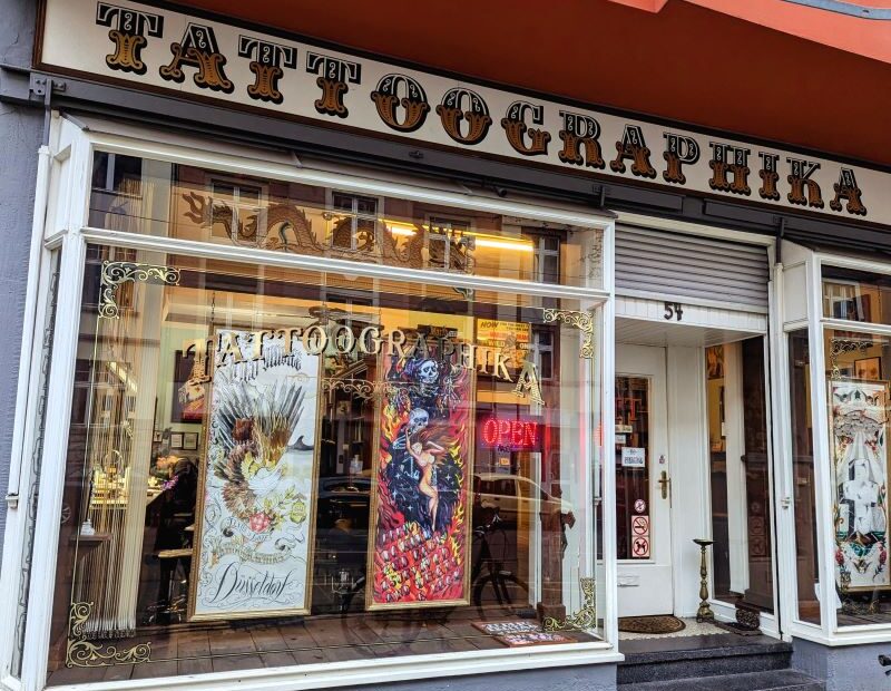 Außenansicht von Tattoographika mit dem ikonischen goldenen Schriftzug und bunten Kunstwerken im Fenster.
